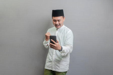 Asian Muslim man wearing a koko shirt and peci with shades of the fasting month, clenching his fist and holding his smartphone while looking to smartphone, isolated on a gray backgroundの写真素材