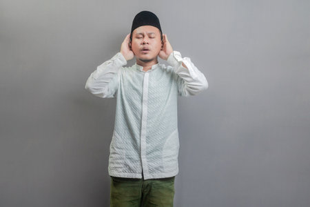 Asian Muslim man wearing a koko shirt and peci with shades of the fasting month, standing reciting the call to prayer, isolated on a gray backgroundの写真素材