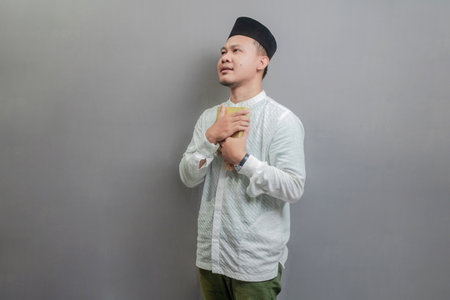 Asian Muslim man wearing a koko shirt and peci with shades of the fasting month, holding prayer beads and reading Qur'an, isolated on a gray backgroundの写真素材