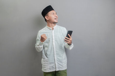 Asian Muslim man wearing a koko shirt and peci with shades of the fasting month, clenching his fist and holding his smartphone while looking at an empty space, isolated on a gray backgroundの写真素材