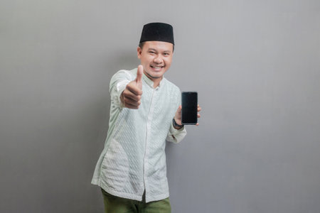 Portrait of an Asian Muslim man wearing a koko shirt and peci with shades of the fasting month, smiling with showing thumbs up and his smartphone, isolated on a gray backgroundの写真素材