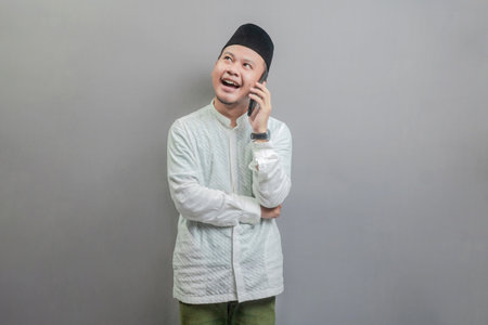 Portrait of an Asian Muslim man wearing a koko shirt and peci with shades of the fasting month, calling with family or friend, isolated on a gray backgroundの写真素材