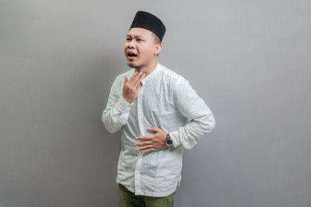 Asian Muslim man wearing a koko shirt and peci with shades of the fasting month, holding back hunger and waiting for the time to break the fast, isolated on a gray backgroundの写真素材