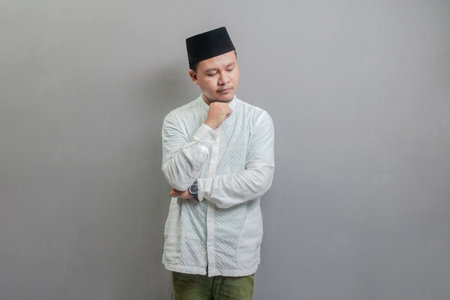 Happy asian muslim man hinking worried about a question, concerned and nervous with hand on chin wearing a koko shirt and peci with shades of the fasting month, isolated on a gray backgroundの写真素材