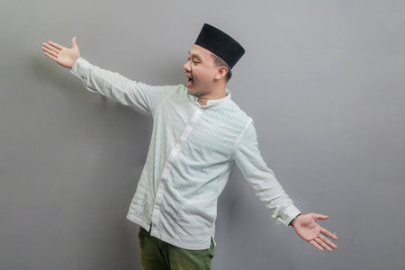 Happy asian muslim man recommend promo as pointing to copy space, indicate down link wearing a koko shirt and peci with shades of the fasting month, isolated on a gray backgroundの写真素材