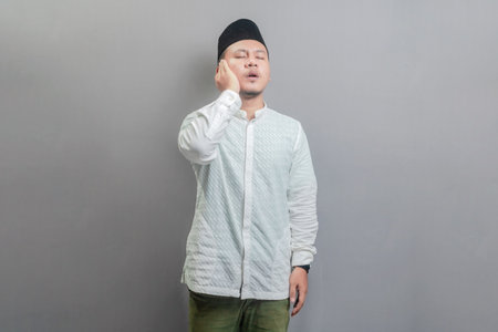 Asian Muslim man wearing a koko shirt and peci with shades of the fasting month, standing reciting the call to prayer, isolated on a gray backgroundの写真素材
