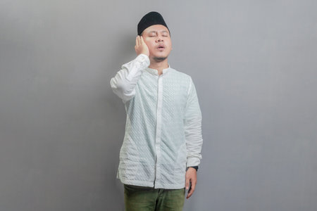 Asian Muslim man wearing a koko shirt and peci with shades of the fasting month, standing reciting the call to prayer, isolated on a gray backgroundの写真素材