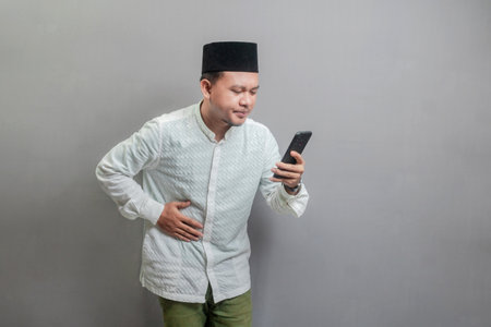 Asian Muslim man wearing a koko shirt and peci with shades of the fasting month, holding back hunger and waiting for the time to break the fast, isolated on a gray backgroundの写真素材