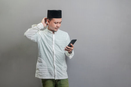 Asian Muslim man standing scratching his head and looking the smartphone, wearing a koko shirt and peci with shades of the fasting month, isolated on a gray backgroundの写真素材