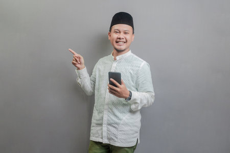 Asian Muslim man wearing a koko shirt and peci with shades of the fasting month, smilling while ponting to empty space while holding smartphone, isolated on a gray backgroundの写真素材