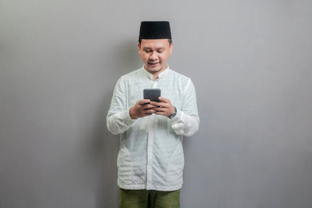 Happy asian muslim man using smart phone cellphone for calls, social media, mobile application online, wearing a koko shirt and peci with shades of the fasting month, isolated on a gray backgroundの写真素材