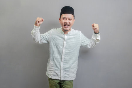 Happy asian muslim man fun successful raised hands show muscles wearing a koko shirt and peci with shades of the fasting month, isolated on a gray backgroundの写真素材