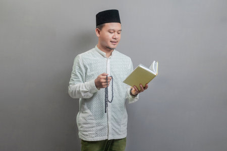 Asian Muslim man wearing a koko shirt and peci with shades of the fasting month, holding prayer beads and reading Qur'an, isolated on a gray backgroundの写真素材