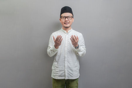 Asian Muslim man wearing a koko shirt and peci with shades of the fasting month, standing looking empty space happily to show grateful , isolated on a gray backgroundの写真素材