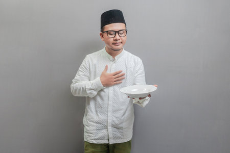 Asian Muslim man wearing a koko shirt and peci with shades of the fasting month, holding a plate while waiting for the time to break the fast, isolated on a gray backgroundの写真素材