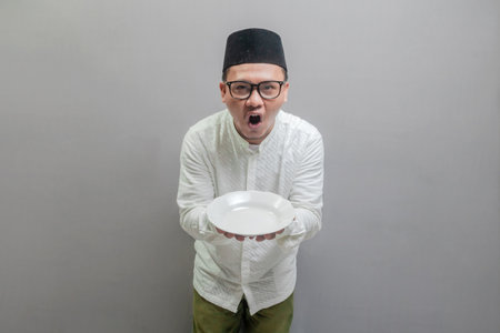 Happy asian muslim man holding empty white plate or dish wearing a koko shirt and peci with shades of the fasting month, isolated on a gray backgroundの写真素材