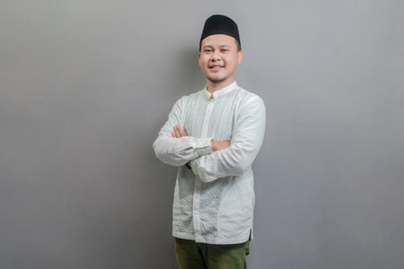 Asian Muslim man standing with crossed arm, wearing a koko shirt and peci with shades of the fasting month, isolated on a gray backgroundの写真素材