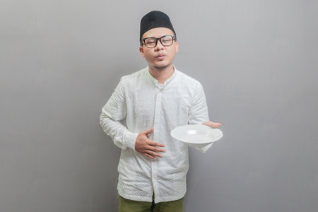Asian Muslim man wearing a koko shirt and peci with shades of the fasting month, holding back hunger and waiting for the time to break the fast, isolated on a gray backgroundの写真素材