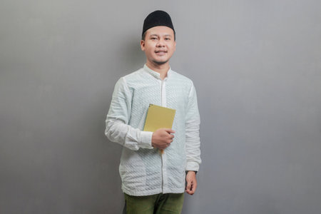 Asian Muslim man wearing a koko shirt and peci with shades of the fasting month, holding prayer beads and reading Qur'an, isolated on a gray backgroundの写真素材