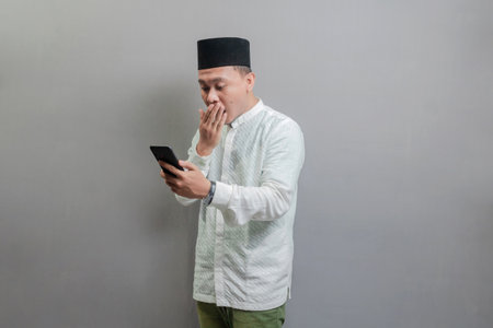 Surprised asian muslim man using smart phone cellphone for calls, social media, mobile application online, wearing a koko shirt and peci with shades of the fasting month, isolated on a gray backgroundの写真素材
