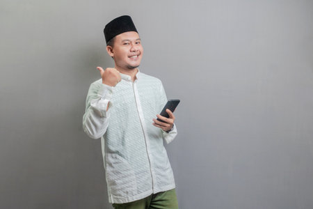 Asian Muslim man wearing a koko shirt and peci with shades of the fasting month, smilling and showing thumbs up and holding smartphone, isolated on a gray backgroundの写真素材