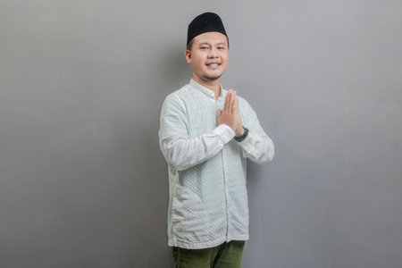 Asian Muslim man wearing a koko shirt and peci with shades of the fasting month, standing with Eid greeting gesture and welcoming Ramadan, isolated on a gray backgroundの写真素材
