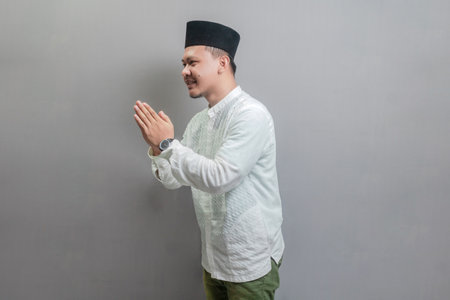 Asian Muslim man wearing a koko shirt and peci with shades of the fasting month, standing with Eid greeting gesture and welcoming Ramadan, isolated on a gray backgroundの写真素材