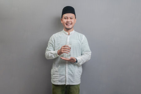 Happy asian muslim man holding glass of pure water give to camera wearing a koko shirt and peci with shades of the fasting month, isolated on a gray backgroundの写真素材