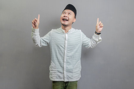 Happy asian muslim man recommend promo as pointing to copy space, indicate down link wearing a koko shirt and peci with shades of the fasting month, isolated on a gray backgroundの写真素材