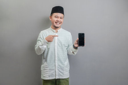 Portrait of an Asian Muslim man wearing a koko shirt and peci with shades of the fasting month, smiling pointing finger to his smartphone, isolated on a gray backgroundの写真素材