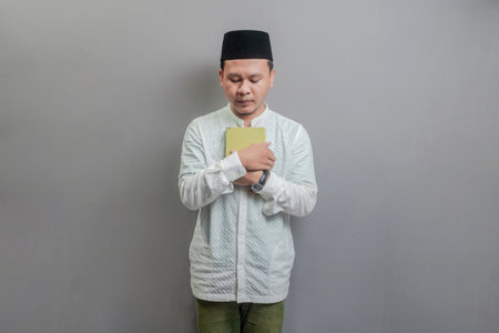 Asian Muslim man wearing a koko shirt and peci with shades of the fasting month, holding prayer beads and reading Qur'an, isolated on a gray backgroundの写真素材
