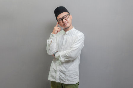 Asian Muslim man wearing a koko shirt and peci with shades of the fasting month, standing doing thinking gesture, looking for idea, isolated on a gray backgroundの写真素材