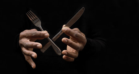 Man's hands holding a knife and fork on a black background.
