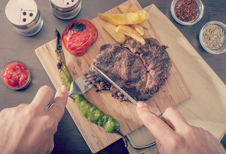 Man cutting and eating a grilled beef steak served with grilled tomatoes, peppers and chipsの写真素材