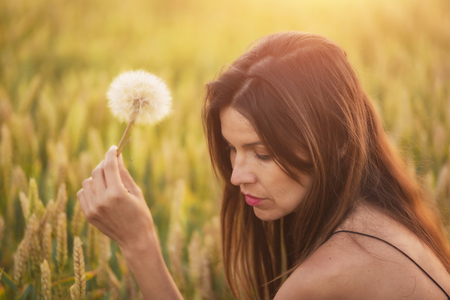 Beautiful young woman blows dandelion in a wheat field in the summer sunset. Beauty and summer conceptの写真素材