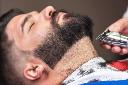 Barber shaving the beard of a handsome bearded man with an electric razor at the barber shop.の写真素材
