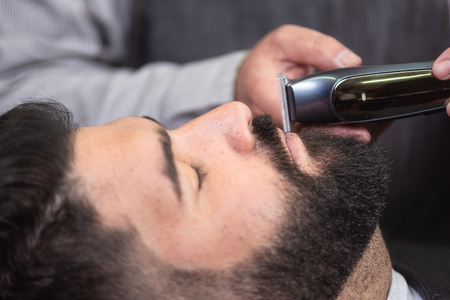 Barber shaving the beard of a handsome bearded man with an electric razor at the barber shop.の写真素材