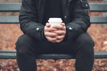 Young man sitted in a bench holding disposable coffee cup in the park in autumn season.の写真素材