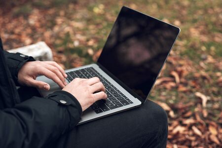 Young man sitting on park bench on autumn with laptop. Student using computer outdoors. Close up.の写真素材