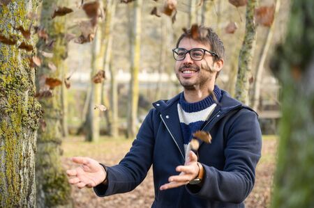 Happy young man playful throwing leaves with open arms dancing under a rain of leaves in autumn...
