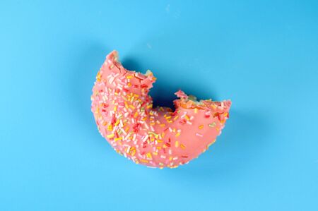 Pink frosted donut with colorful sprinkles with bite missing. On blue background.