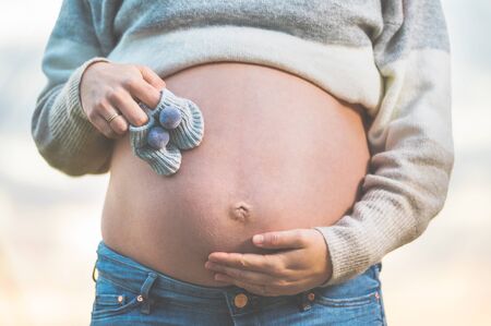 Pregnant woman posing outdoors holding little baby shoes on her belly.