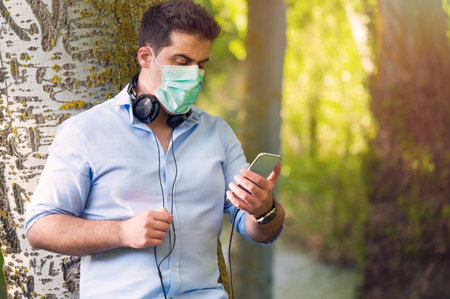Young Man with protective face mask listening to music outdoors in the park while maintaining social distance. Lifestyle during Covid-19 pandemic outbreak.の写真素材