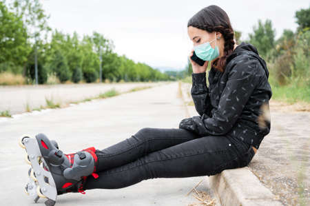 Woman in protective face mask on roller skating pause, sitting on the street and using mobile phone...