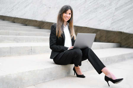 Beautiful businesswoman in suit, using a laptop sitting on stairs outdoors