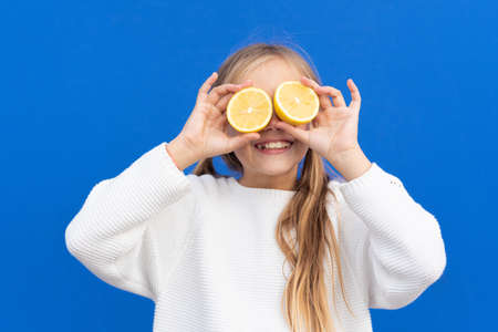 Overjoyed blond haired girl in white sweater smiling while covering eyes with bright lemon slices isolated on blue backgroundの写真素材