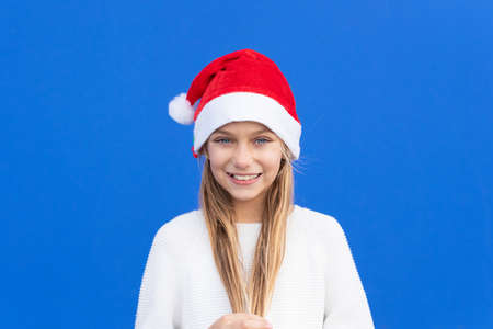 Pleased girl in white sweater and Santa hat holding pigtails smiling away isolated on blue backgroundの写真素材