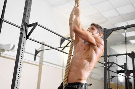 Fitness man doing rope climb exercise in gym.