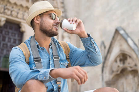 Happy young tourist man with coffee to go and backpack resting on street.の写真素材
