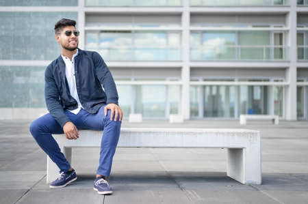Portrait of young man in stylish sunglasses posing while sitting in bench in urban background.